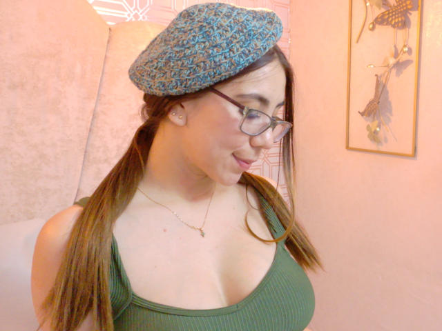 FreyaraVenwo with glasses, green ribbed tank top, blue patterned flat cap, pigtails, fair skin, small earrings, subtle smile, medium boobs, necklace, framed art on pink wall.