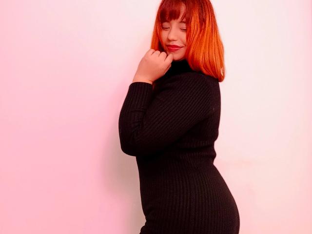 BelleVivienneHt con un corte de pelo bob rojo-anaranjado, vistiendo un cuello alto de punto negro, con los ojos cerrados, sonriendo, con la mano cerca de la barbilla, de pie de lado, resaltando sus curvas y sus pechos medianos..