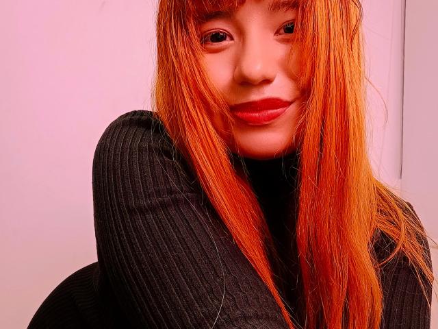 BelleVivienneHt with red-orange hair, wearing a black ribbed turtleneck, smiling with red lipstick, her tits covered by her arm, pinkish light on her face.