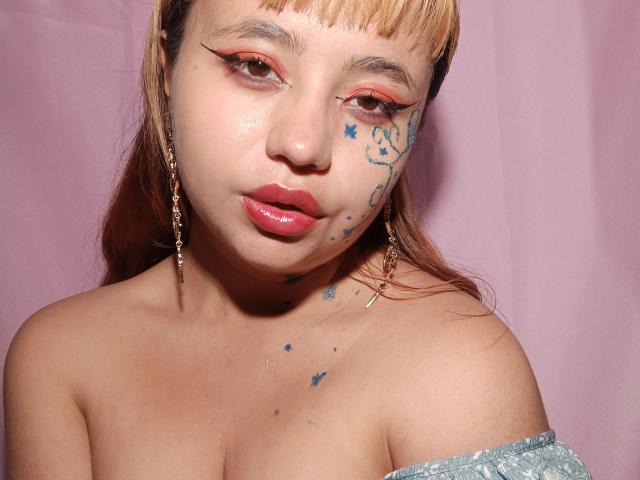 BelleVivienneHt with light brown hair, red eyeliner, blue star tattoos on her face and chest, pink lips, and off-shoulder blue top, wearing gold earrings, showing her tits.
