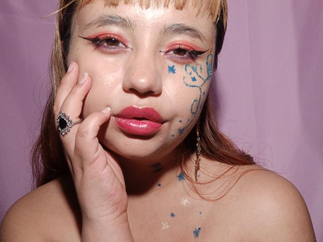 BelleVivienneHt has shiny, pink-lipsticked pout, red eyeshadow, and blue star tattoos on her face. She touches her cheek with her hand, wearing a jeweled ring.