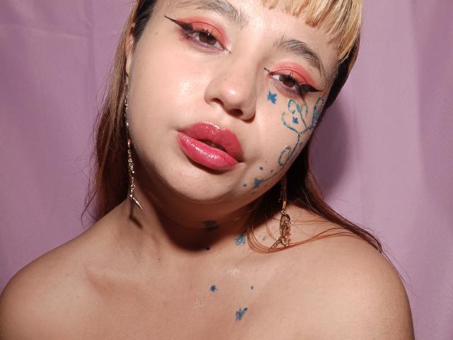 BelleVivienneHt with pink lipstick, star tattoos on cheek, and brown hair, looking slightly to the side, has shiny skin and wears chain earrings. Boobs not visible.