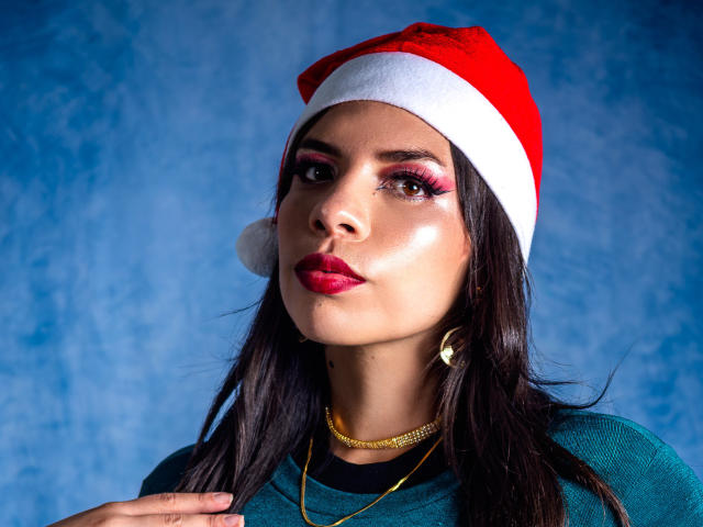 VioletaStone with red Santa hat, pink eye makeup, red lipstick, green shirt, gold necklace, and earrings, has dark hair and a serious expression. Blue textured background.