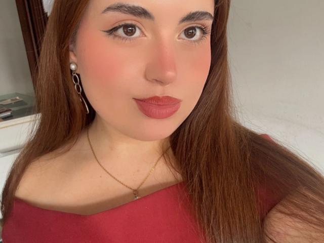 VicluX with long brown hair, pink blush, winged eyeliner, red lipstick, pearl earrings, gold necklace, and a red top, smiling at the camera.
