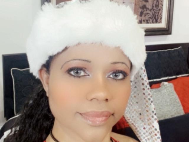Cincia69 hat eine hellbraune Haut, schwarzes, lockiges Haar und trägt einen flauschigen, weißen Weihnachtsmannhut. Sie lächelt mit rosafarbenen Lippen und trägt dezentes Make-up mit rosafarbenem Lidschatten..