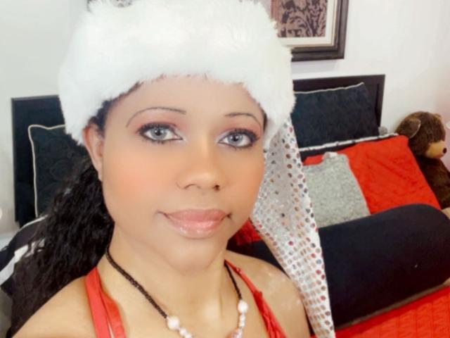 Cincia69, com pele marrom média, cabelo preto encaracolado, usando um chapéu branco de pele, top vermelho de alça, colar de pérolas e maquiagem discreta, sorrindo para a câmera..