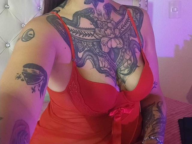 AmberKeller with large tattoos covering her chest and arms, wearing a red lingerie dress with a bow, showing her tits and a snake tattoo over her cleavage.