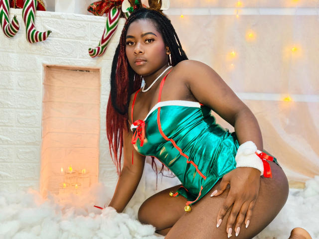 KarinaFlowers with dark skin and red-dyed braids, wearing a shiny green Christmas outfit, red ribbon, and white fur, kneeling on white fluff, with bells on her thigh.