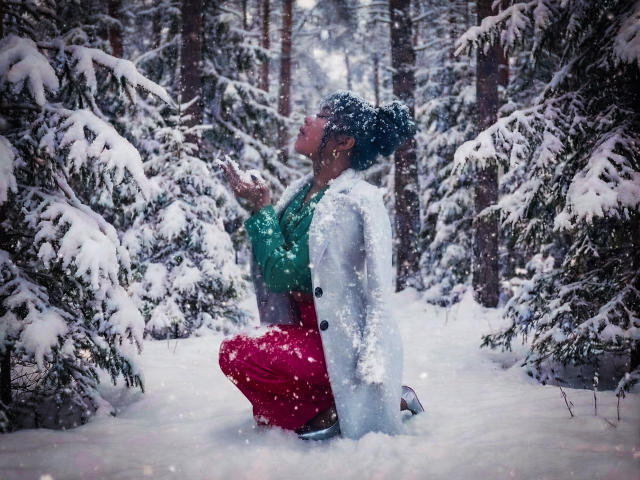 SophieSonyas kneeling in snow, wearing a green top, red pants, and grey coat, snowflakes on face, black curly hair, forest background, snowy trees, mid-action, winter scene.