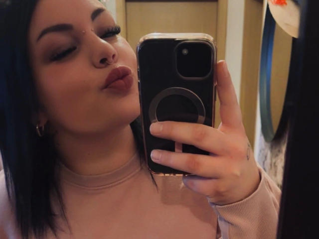 MadameMarie-hot with black hair, pouty lips, and nose ring, taking a selfie with a black phone, wearing a pink top, in a mirror reflection.