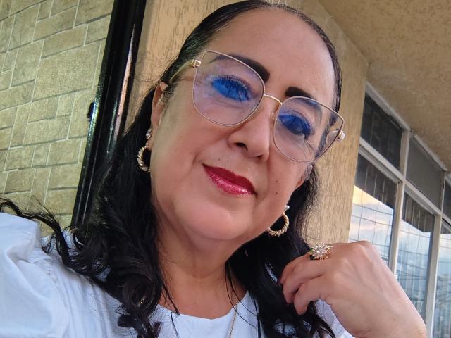 Amarantac with blue eyeshadow, red lipstick, glasses, black hair, white shirt, gold earrings, ring, and hoop earring, smiling at camera. Her pussy's not visible.