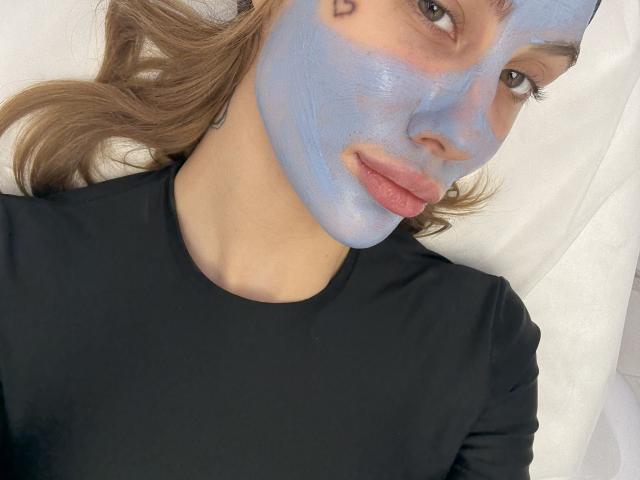 SensualMila with blue face mask, wavy brown hair, black shirt, and pink lips, looking at camera with small heart tattoo above right eye.