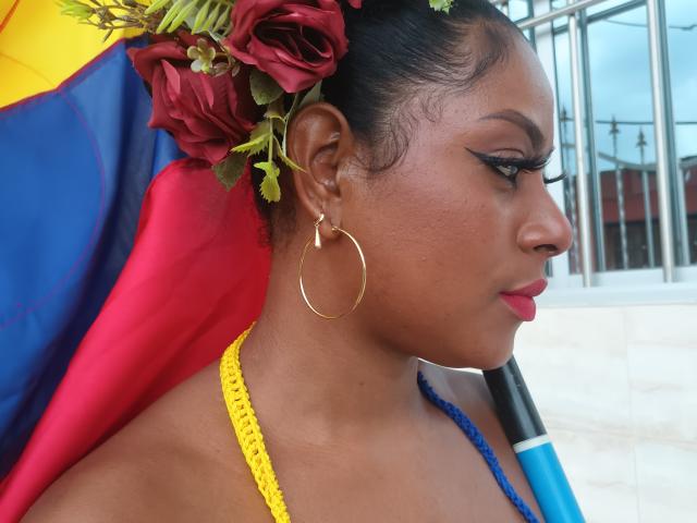 PamelaLce has dark skin, red roses in her hair, gold hoop earrings, yellow necklace, blue strap, and dramatic eyeliner. Her profile shows her nose and pink lips.