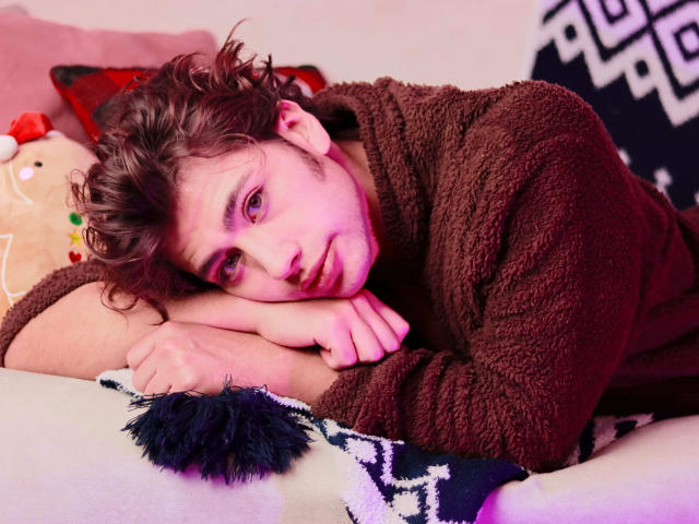 TonyDan with curly brown hair, purple eyes, and a fluffy brown sweater, lying on a pillow, head resting on his hand, looking tired and sexy. Soft pink light.