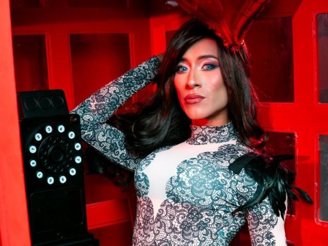 ThomasParker69 with blue eyes, wearing a lace dress, black feather accessory, and red light, has long brown hair, and a confident expression, standing next to a red wall.