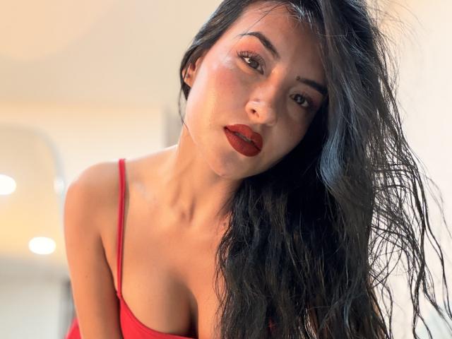 EmilyJensenn with wet black hair, red lipstick, and red tank top, showing her tits and looking down with a slight smile. Soft, natural light on her face.