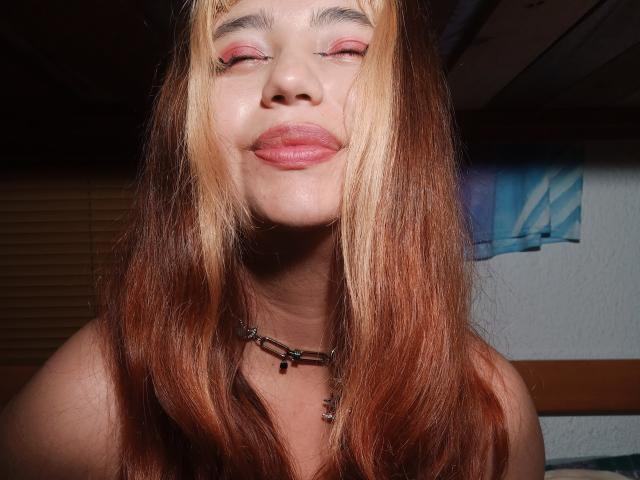 BelleVivienneHt with closed eyes, pink lipstick, and long brown-to-blond ombre hair, smiling softly. She's wearing a necklace with small beads. Her skin is light.