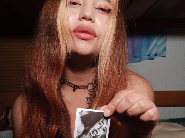 BelleVivienneHt with long brown and blonde hair, pink lipstick, and a chain necklace, holding a black-and-white photo of a woman's face, pointing at it.