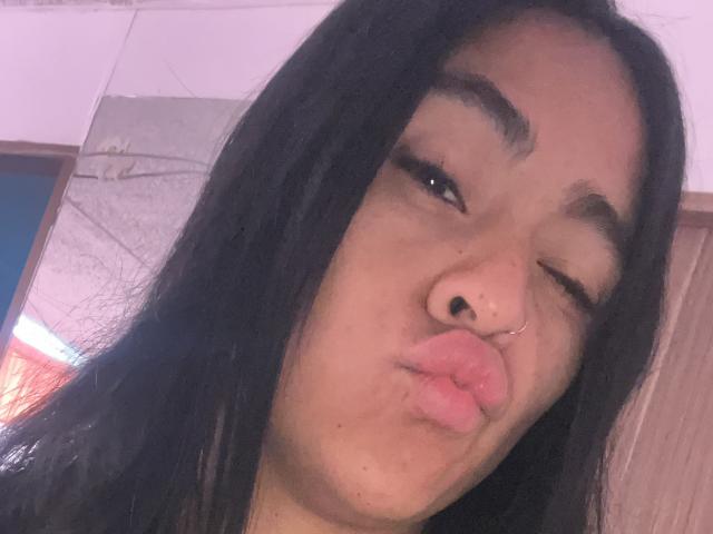 StellaBeckker has black hair, a nose ring, and is pouting her pink lips with one eye closed. She's looking cute and has a small black nose dot.