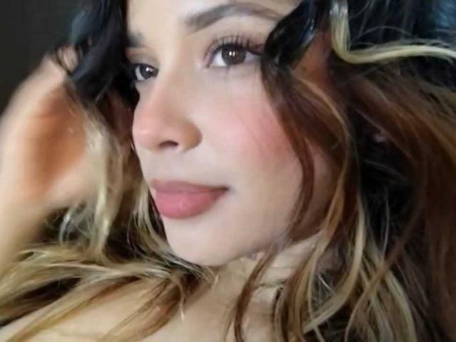 KateZoe's face, showing brown eyes, curled black and blonde hair, pink lips, and a slight smile. Her hand is near her head, with a soft, warm light.