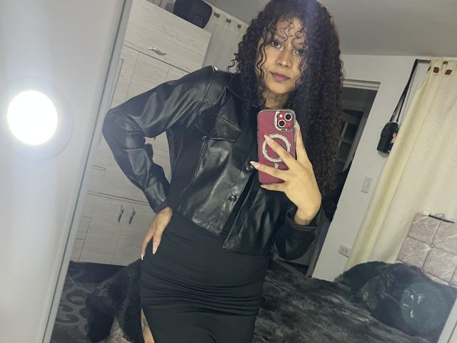 Sofia, met krullend bruin haar, draagt een zwarte leren jas en een strakke zwarte rok, en maakt een selfie in de spiegel met een roze telefoon. Ze poseert zelfverzekerd en heeft een lichte glimlach..