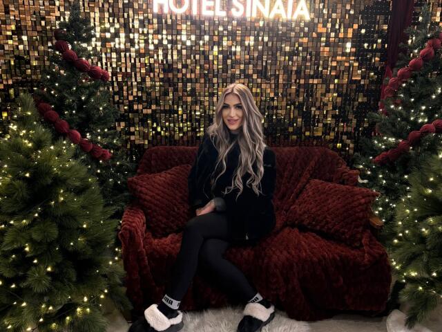 NicolSNaya with long blonde hair, black sweater, black leggings, and white sock shoes, sitting on a red velvet couch between two Christmas trees with lights.
