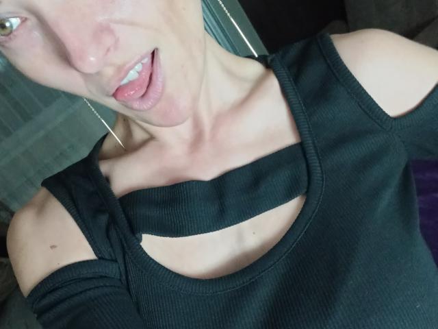 BellaJulls with a tongue out, wearing a black ribbed top with off-shoulder straps, showing collarbone and small moles, earrings visible, light skin, short hair, slightly open mouth.