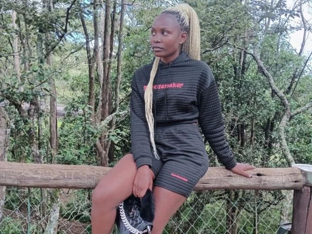 SexxySunflower-hot with long blonde braids sits on a wooden fence, wearing a dark gray waffle dress, showing her thighs, with black sneakers in her hand.