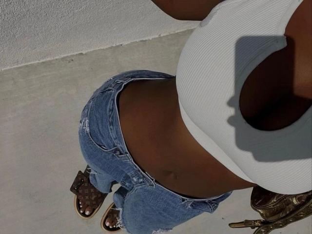 SexxySunflower-hot's boobs in a white ribbed tank top, showing a tan belly, wearing frayed blue jeans, brown croc sandals, with a shadow of her face on the right.