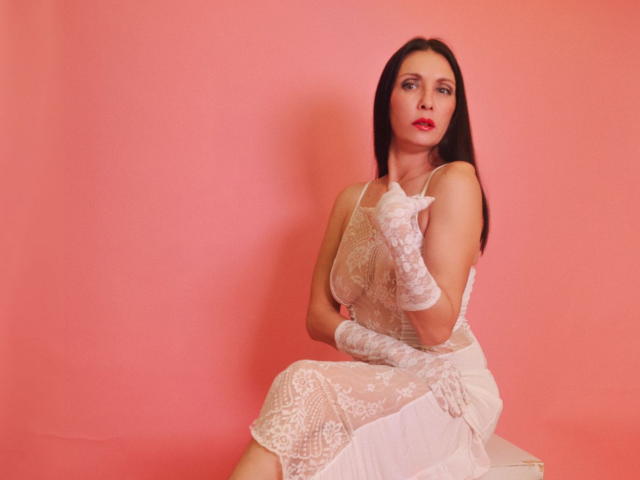 EvanessaJohn with pale skin, dark straight hair, red lipstick, white lace dress, and matching gloves, sitting on a box against a pink wall, looking seductively at the camera.