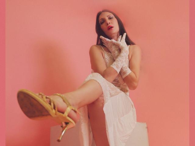 EvanessaJohn has long brown hair, wearing a white lace dress and gloves, with gold strappy heels, exposing her pussy under the dress, against a pink wall.