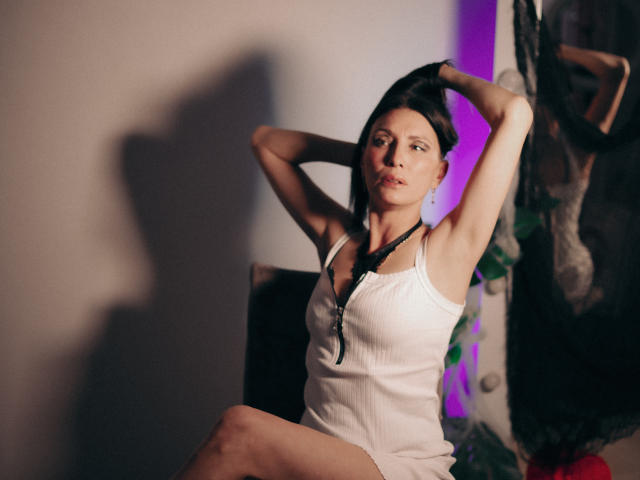 EvanessaJohn with arms raised, wearing a white ribbed tank top, exposing her small tits and a black necklace, sitting with a serious expression in a dimly lit room.