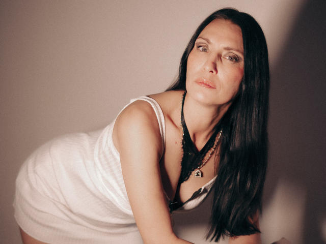 EvanessaJohn with long black hair, wearing a white dress with black lace, leaning forward, showing her tits, wearing a black necklace, in a soft light, looking at camera.