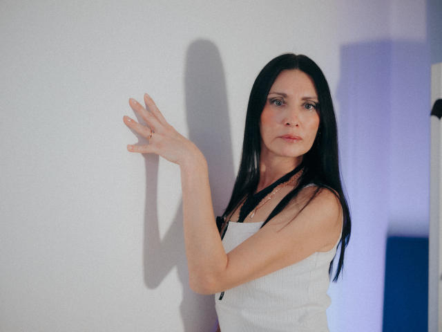 EvanessaJohn with black hair, white tank top, and hand against wall, showing off her tits, pale skin, and a subtle smile in a well-lit, minimalist setting.