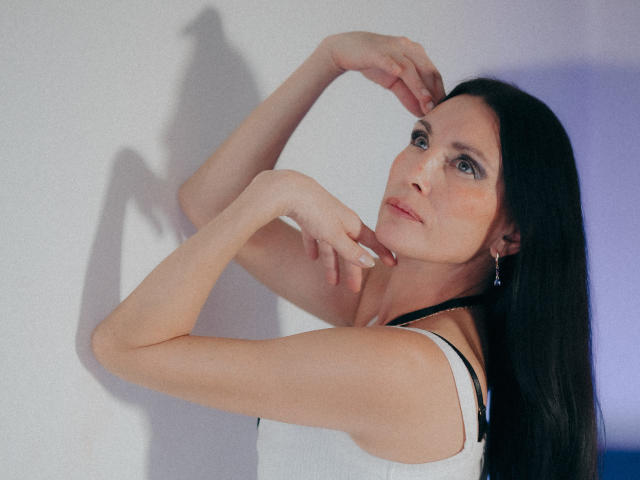 EvanessaJohn with long black hair, pale skin, and blue eyes, wearing a white tank top, has her hands near her face, showing off her small tits and delicate earrings.