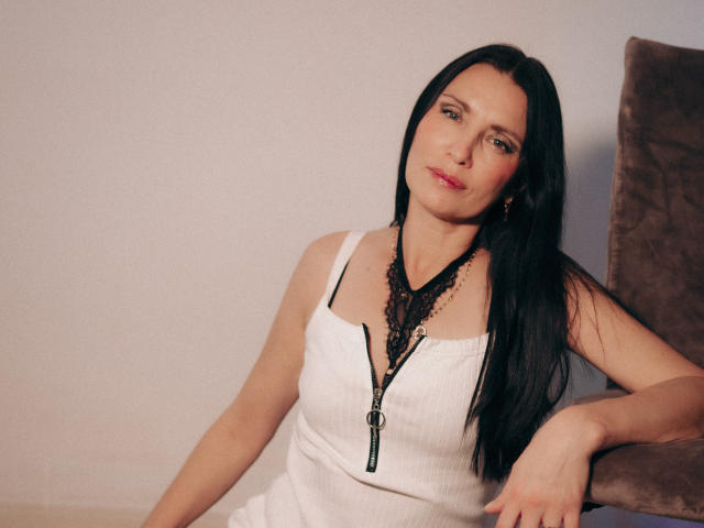 EvanessaJohn with long black hair, wearing a white zip-up dress, black lace necklace, and red lipstick, sitting on a brown chair, looking slightly to the side.