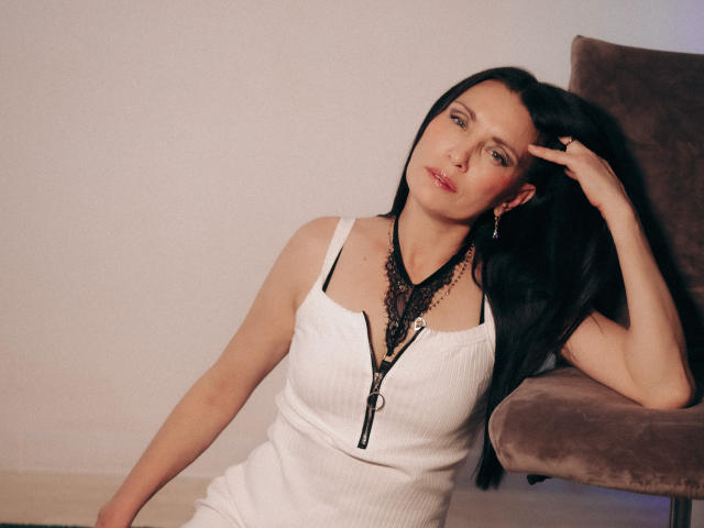 EvanessaJohn with long black hair, white ribbed dress, black lace necklace, and earrings, leaning on a brown chair, hand in hair, red lipstick, and a slightly seductive look.