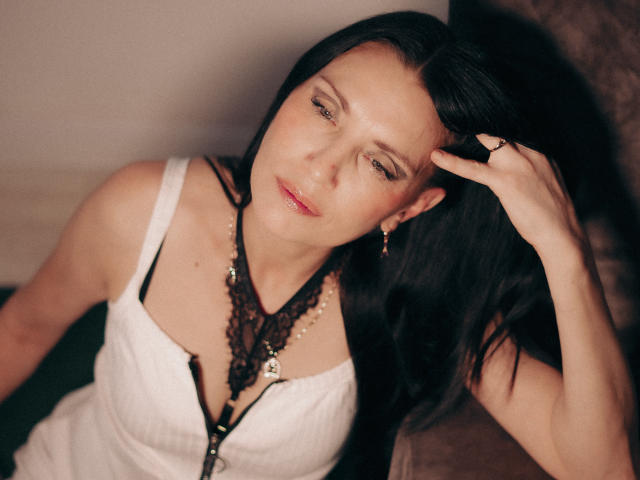 EvanessaJohn with long black hair, wearing a white tank top, black lace necklace, and gold earrings, touches her head with a ringed hand, looking slightly down.