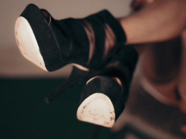 EvanessaJohn's black high heels with white soles, pointed toes, and straps are in focus, showing worn-out bottoms and her ankles. Her legs are slightly crossed.