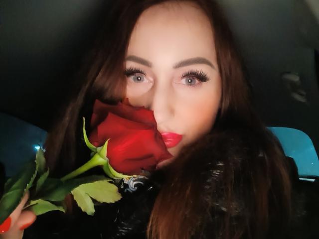 FreyaHott69 mit einer roten Rose im Mund, die den Stiel mit grünen Blättern hält, dunkles braunes Haar, starker Eyeliner, lange Wimpern, leuchtend roter Lippenstift, schwarzer Fellmantel, blaue Autositzlehne..