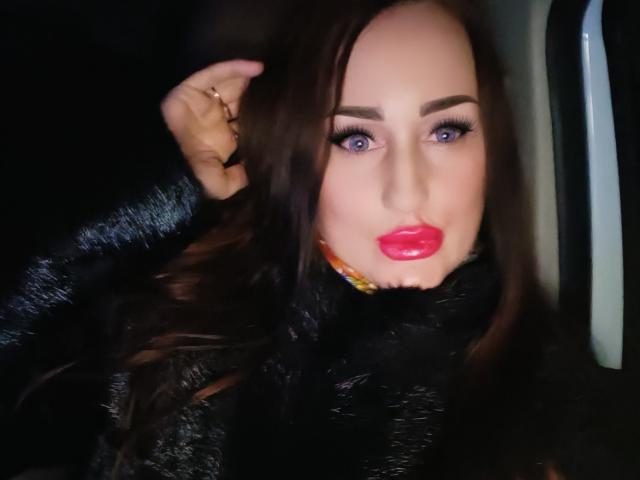 Close-up of FreyaHott69 with light skin and long dark brown hair, wearing bright red lipstick and dark eye makeup with thick eyeliner. She has a gold ring on her right hand, slightly raised to her hair. She's dressed in a black, textured coat. The image has a slightly blurred effect, with her face centered and a dark, out-of-focus background. Her expression is neutral, and the lighting highlights her facial features and lipstick.