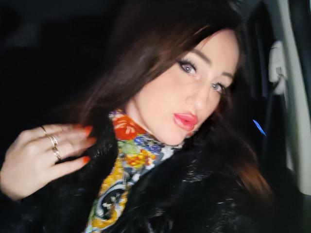 FreyaHott69 with dark brown hair, red lipstick, and heavy makeup, wearing a black fur coat and colorful scarf, showing off orange nails and gold rings, looking sexy.