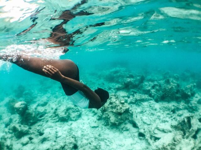 Underwater photograph of KharlaQueen. She's floating horizontally, her dark skin contrasting with the clear blue water. Her arms are folded in front of her chest, and she's wearing a white swimsuit. The water's surface ripples above her, and the rocky seabed is visible in the background. Sunlight filters through, creating subtle shadows on her body. The image uses natural light, highlighting the underwater environment's vibrant blue hues and the texture of the rocks. KharlaQueen's reflection is faintly visible in the water's surface.
