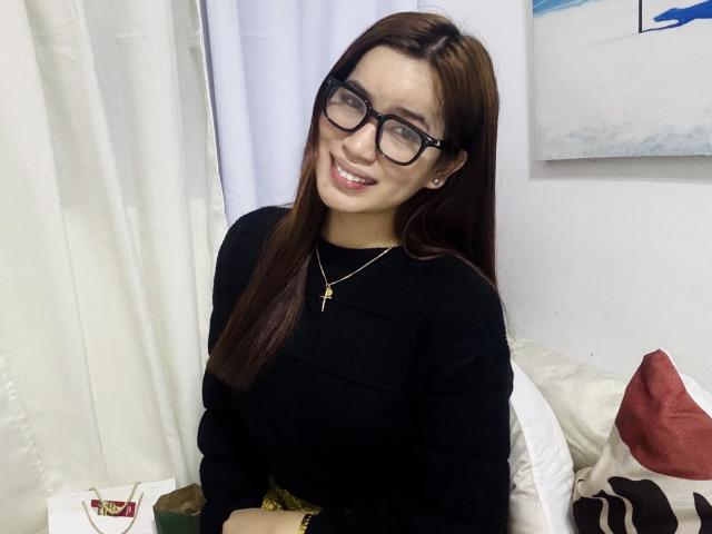DaphneyLove with long brown hair, black glasses, and a black long-sleeve top, smiling at the camera, wearing a gold cross necklace, sitting on a white couch.