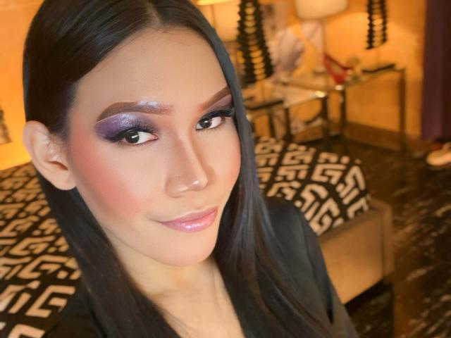 KellyShemale has long black hair, a smooth face, and glossy pink lips. She wears shimmering purple eyeshadow, dark eyebrows, and a subtle blush. Her dark eyes look slightly to the side. She's dressed in a black top. The background is blurred, showing a geometric-patterned black and white blanket on a bed and a lamp. The image has a warm, yellowish tone. KellyShemale's makeup is bold and well-applied, contrasting with her natural skin tone.