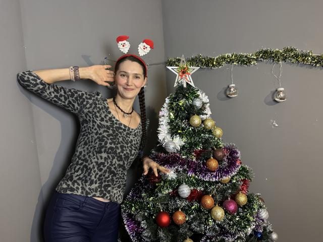 TayaFlavy with red Santa ears, leopard print shirt, and bracelet, stands next to a decorated Christmas tree with glitter, ornaments, and a star topper. Her ass is visible.