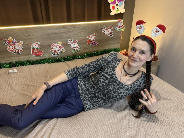 TayaFlavy with long brown braid, wearing leopard print top, blue pants, red headband with bunny ears, necklace, and bracelet, lying on bed, smiling, in Christmas-themed photo.