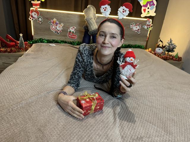 TayaFlavy is lying on a beige bed with a textured cover, wearing a leopard print shirt and holding a small, red gift box with a gold ribbon in her right hand. She holds a plush Christmas figure with a red hat and white face in her left hand. Her dark hair is pulled back with a red headband. Christmas decorations, including a Santa figure, wreaths, and ornaments, are on the wooden headboard behind her. She has a light skin tone, blue eyes, and a subtle smile. She is wearing a black choker necklace and has a tattoo on her right wrist.