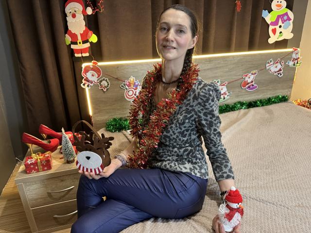 TayaFlavy with brown hair, wearing leopard print shirt, red and green tinsel, blue pants, holding reindeer ornament, sitting on bed with Christmas decor, wooden drawer, and red heels.