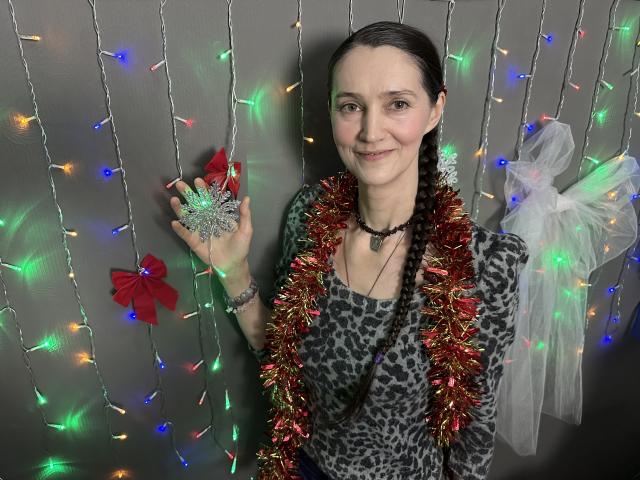 TayaFlavy stands in front of a grey wall adorned with colorful, glowing Christmas lights. She has a fair complexion, dark braided hair, and a subtle smile. She wears a leopard-print shirt, a red and gold tinsel garland around her neck, and holds a glittery heart-shaped ornament with a red bow. A white, tulle-winged accessory is attached to her back. Her necklace and bracelet add subtle details. The overall composition is festive and slightly quirky.
